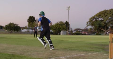 Cricket batsman sprinting to crease at dusk driving bat to complete run on suburban pitch