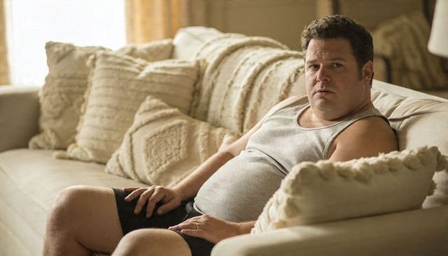 Middle-aged man reclining on beige sofa wearing gray tank top and wedding ring