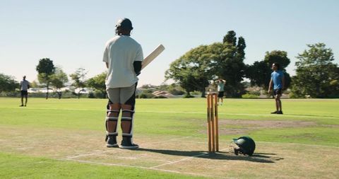 Cricket batsman standing on marked pitch holding bat beside stumps and spare helmet