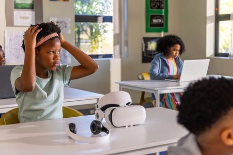 African american girl adjusting headband by vr headset while classmates using laptops