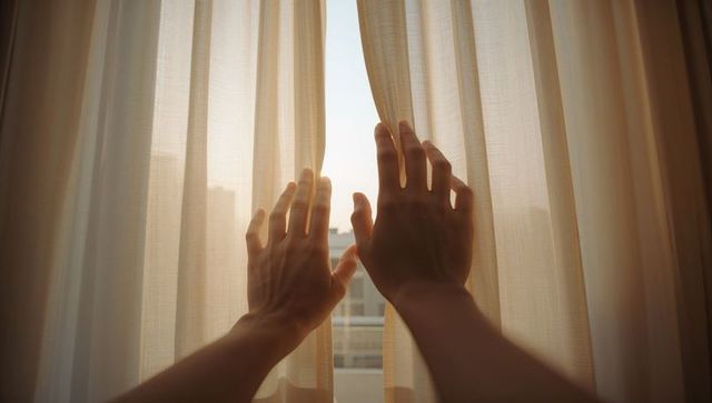 Parting sheer curtains revealing soft morning sky over city, hands and forearms reaching