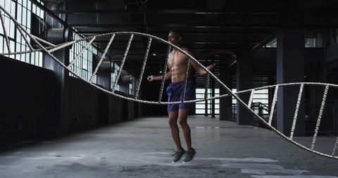 Shirtless male athlete jumping rope in abandoned industrial warehouse, moody fitness shot