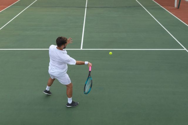Rear view of Caucasian man wearing tennis whites spending time on a court playing tennis on a sunny day, holding a tennis racket hitting ball. Hobby sport leisure time.