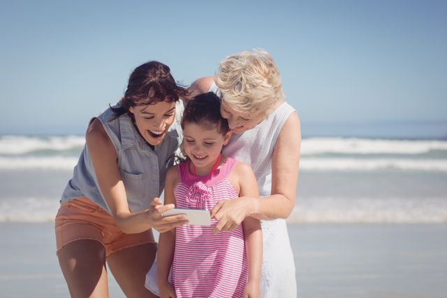 Three generations of women, including a grandmother, mother, and daughter, are enjoying a sunny day at the beach. They are gathered around a mobile phone, smiling and sharing a moment of joy. This image is perfect for promoting family vacations, technology use in everyday life, and the importance of family bonding. It can be used in advertisements, travel brochures, and social media campaigns highlighting family togetherness and outdoor activities.