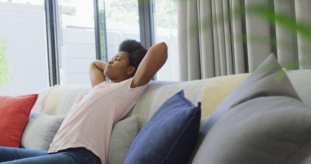 African American woman lounging on sofa, stretching arms behind head. Suitable for themes related to relaxation, home comfort, leisure time, mental wellness, self-care, lazy Sunday.