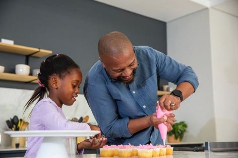 African american father decorating cupcakes with daughter in modern kitchen