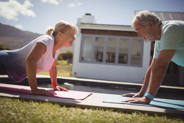 Senior couple performing push-ups on yoga mats near a swimming pool on a sunny day. Both individuals are smiling and appear to be enjoying their workout. This image can be used for promoting senior fitness, healthy lifestyles, outdoor activities, and wellness programs for the elderly.