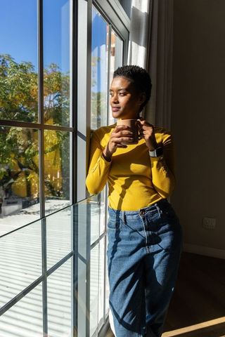 Sunlit morning with african american woman standing by window holding mug in yellow top