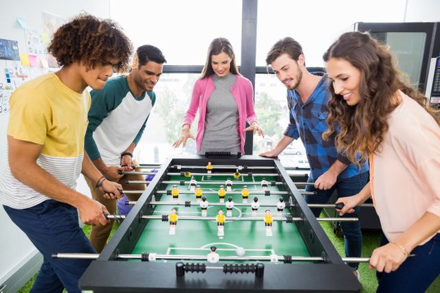 Group of young colleagues enjoying a game of table football in a modern office. They are smiling and interacting, showcasing teamwork and bonding in a casual work environment. Ideal for use in articles or advertisements about workplace culture, team building activities, or promoting a fun and engaging office atmosphere.