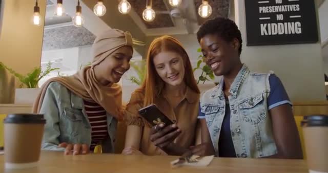 Three young women from diverse backgrounds gathered in coffee shop, sitting together, looking at something on smartphone, smiling and interacting with each other. Useful for promoting community diversity, social activities, technology usage, or coffee shop ambience.