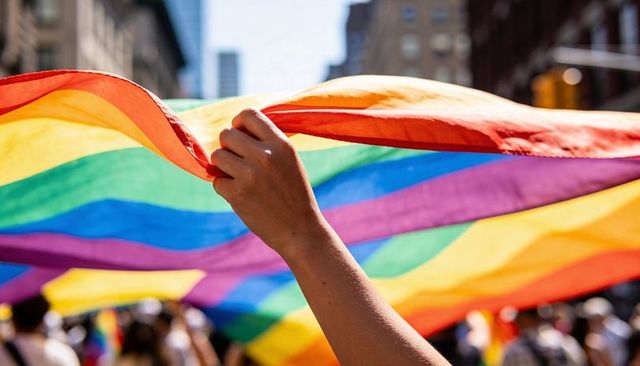 Hand holding giant rainbow pride flag over crowded city street parade, backlit fabric