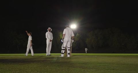 Night cricket match under floodlights with batsman at crease and fielders awaiting delivery