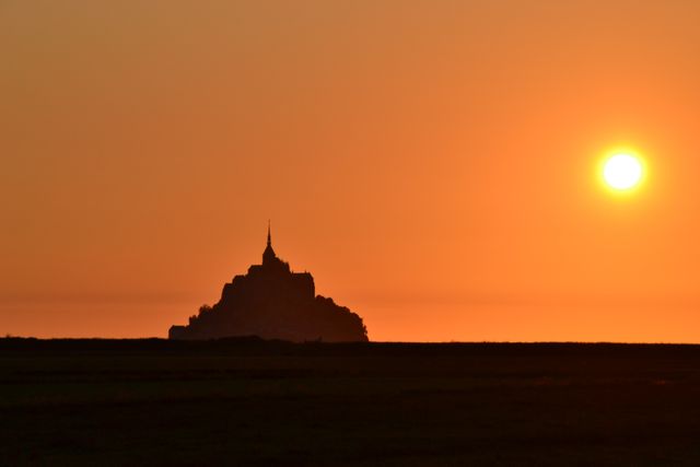 This photo depicts Mont Saint-Michel silhouetted against a vibrant orange sunset, creating a dramatic and serene scene. Mont Saint-Michel is a famous island commune in France, renowned for its medieval architecture and stunning coastal views. This image is great for travel websites, promotional materials, or articles about famous landmarks and historical sites. It can also be used in backgrounds, headers, or posters to evoke a sense of wonder and elegance.