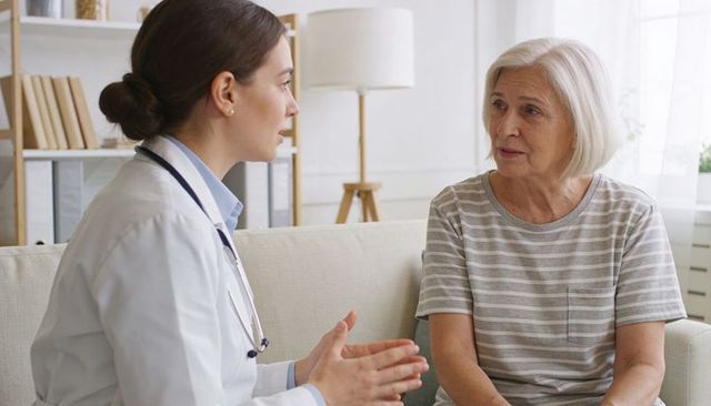 Female doctor consulting elderly woman on home sofa discussing care and treatment options