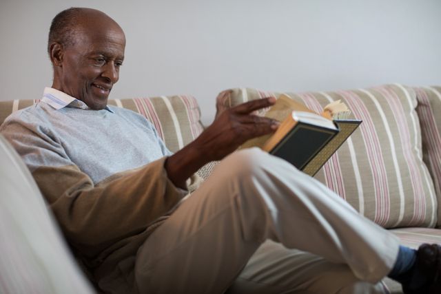 Senior man enjoying a book while sitting comfortably on a sofa at home. Ideal for use in articles or advertisements related to senior living, retirement, leisure activities, and home comfort.