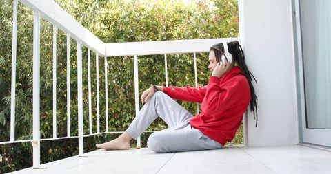 Young man relaxing on balcony listening to music in red hoodie and white headphones