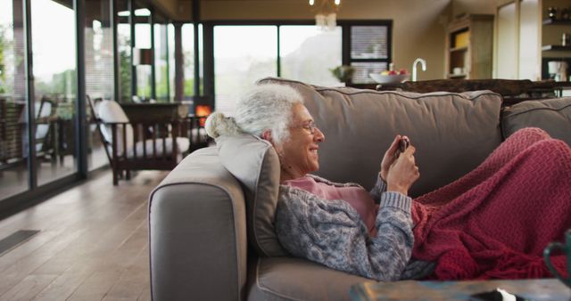 Elderly woman lying on a comfortable couch, using a smartphone. Ideal for depicting an active senior embracing technology, leisure time activities at home, or promoting products for elderly comfort and digital inclusivity.