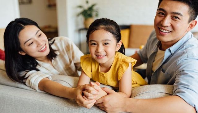 Asian family smiling and holding daughter on cozy sofa in warm living room parental bonding