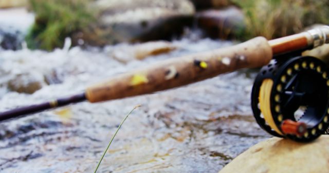 A close-up of a fly fishing rod resting on a rock by a stream, with copy space. The focus on the fishing equipment against the blurred water background suggests a peaceful outdoor activity.