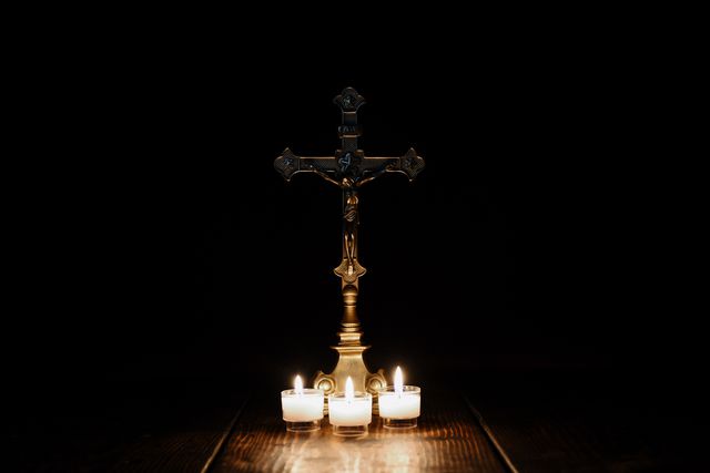 Bronze cross standing with three burning candles on a dark background creating a spiritual and peaceful atmosphere. Ideal for religious occasions, Christian decorations, prayer meditation visuals, and faith-based projects. Can be used in posters, websites, and publications focused on spirituality and Christianity.