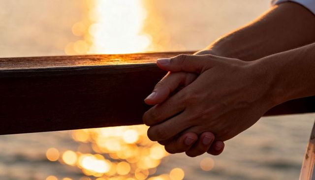 Holding clasped hands resting on wooden railing at sunset with golden water bokeh