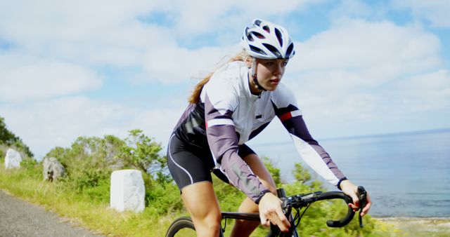 Woman cycling along a scenic coastal road wearing helmet and cycling attire. Ideal for use in campaigns promoting outdoor sports, fitness, active lifestyle, cycling gear, and travel vacations by the coast.