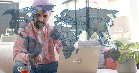 Sikh man working remotely using laptop and phone with global analytics world map overlay