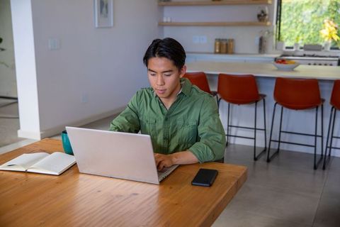 Asian man working on laptop at wooden table in modern open-plan kitchen, home office