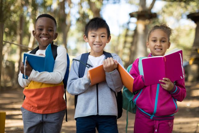 Three diverse children are standing in a park on a sunny day, each holding a book and smiling. They are wearing casual clothing and backpacks, suggesting they are students. This image can be used for educational content, promoting reading, diversity in education, outdoor learning activities, and childhood friendship.