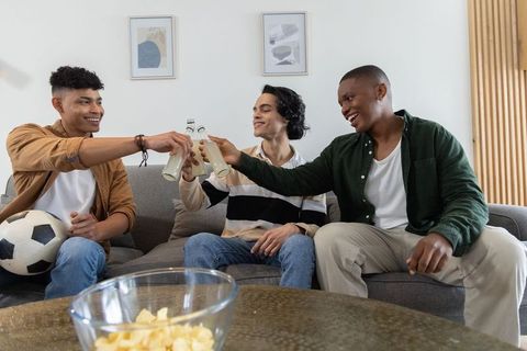 Diverse young men clinking bottles on gray sofa with soccer ball and snacks