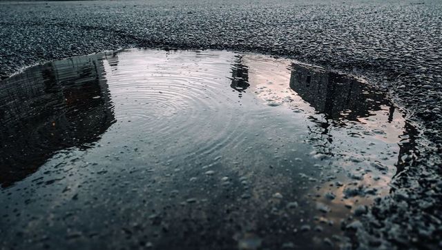 Puddle reflecting city skyline while rippling across wet pebbled asphalt at dusk