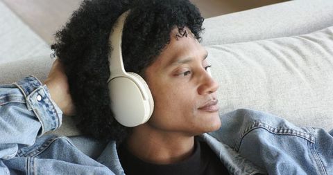 Reclining man listening with white headphones on couch in casual denim jacket at home