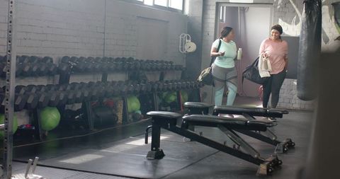 Diverse mid adult women walking into industrial gym carrying duffel bags and water bottle
