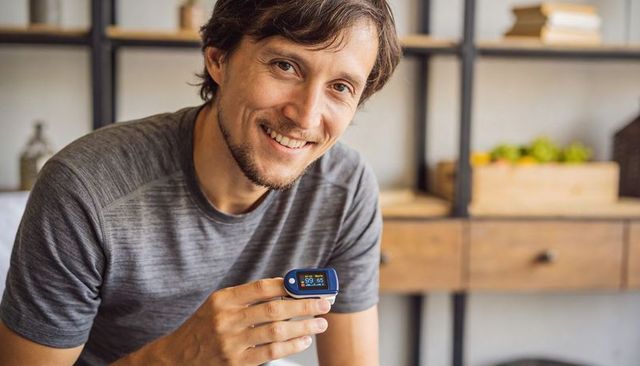 Smiling man holding fingertip pulse oximeter showing spo2 and pulse rate at home