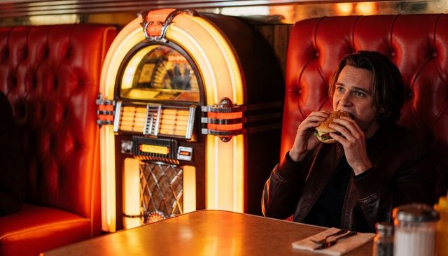 Eating man in leather jacket holding burger at neon-lit 1950s diner booth with jukebox retro