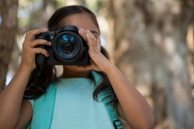 Little girl with pigtails and backpack taking photos with a DSLR camera in a forest. Ideal for themes related to childhood, outdoor activities, photography hobbies, nature exploration, and learning new skills.