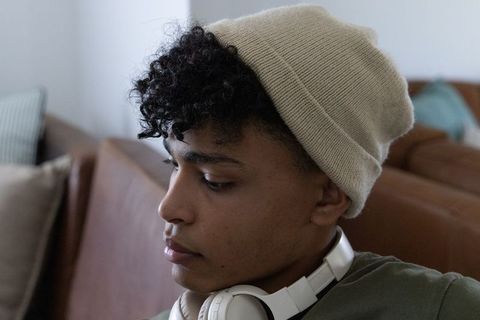 African american man wearing beige beanie and white headphones sitting on brown leather sofa