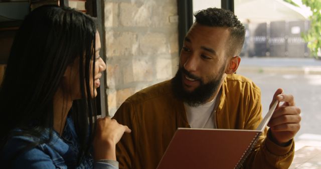 Young couple smiling and discussing something while sitting at a table in a cozy cafe. Man holding a notebook while looking attentively at the woman. Perfect for content related to relationships, communication, casual outings, coffee shops, and intimate conversations.