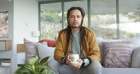 Relaxed man with dreadlocks holding mug on modern sofa in bright contemporary living room