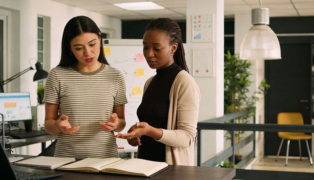 Two women collaborating in modern office discussing strategy and pointing at notebooks