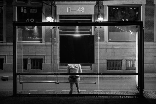 Person sitting alone at a nighttime bus stop illuminated by street lights. The scene depicts quiet urban solitude and public transportation. Ideal for articles or ads related to night photography, urban life, loneliness, and city infrastructure.