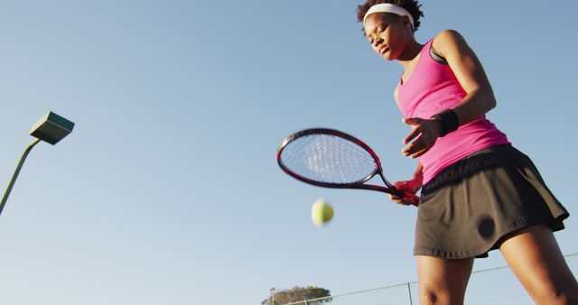 Image of low angle of focused african american female tennis player holding racket and hitting ball. professional tennis training, sport and competition concept.