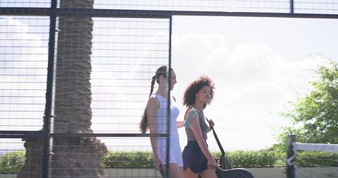 Diverse female friends walking on court in sportswear with racket bag and phone armband