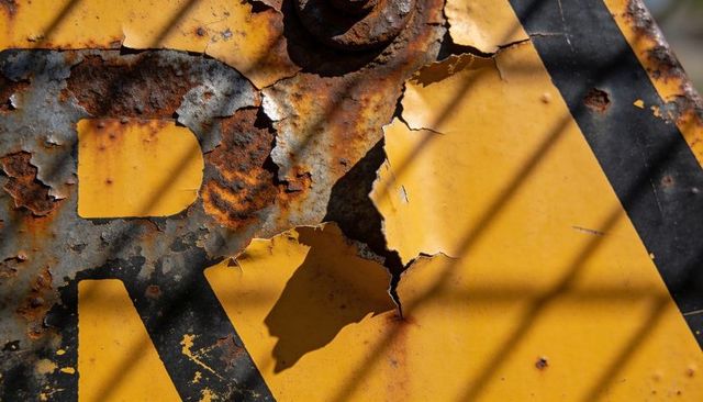 Peeling yellow road sign with rust bolt and fence shadows showing weathered texture
