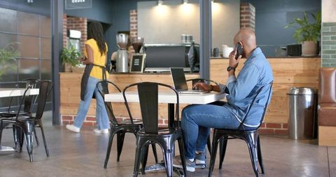 Remote worker making phone call while typing on laptop in modern coffeehouse workspace
