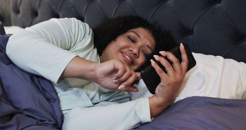 Mid-adult woman reclining in bed using smartphone, scrolling messages in cozy loungewear