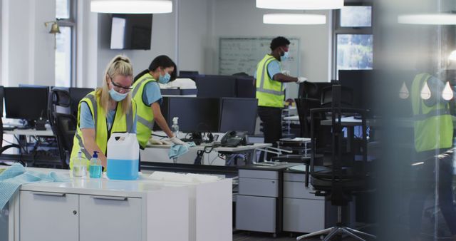 Professional cleaners in high-visibility vests and masks disinfecting an office workspace. Ideal for content related to workplace hygiene, professional cleaning services, and pandemic safety measures.