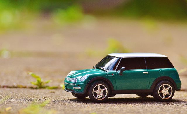 Miniature green car toy is sitting on a pavement outdoors with a blurred natural background. Ideal for use in toy store advertisements, children's playtime themes, automotive enthusiast blogs, and model car showcase materials.