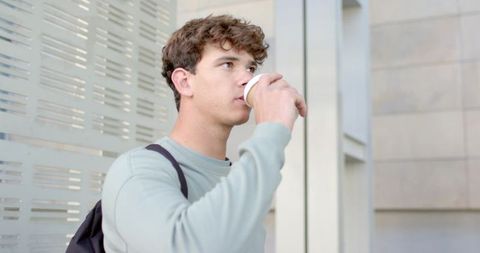 Young man sipping takeaway coffee outside modern office building with backpack on shoulder
