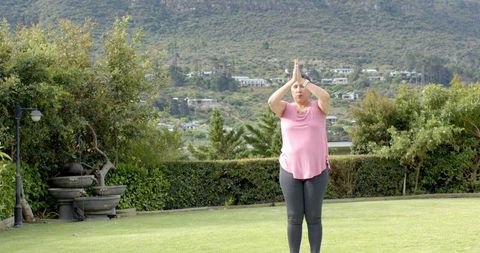 Mature woman practicing yoga in backyard garden pressing palms overhead wearing pink top